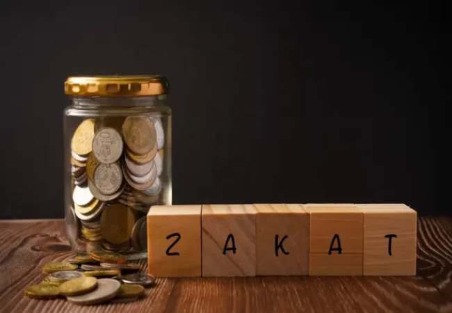 A glass jar filled with coins alongside wooden blocks spelling out the word 'zakat' against a dark background in short