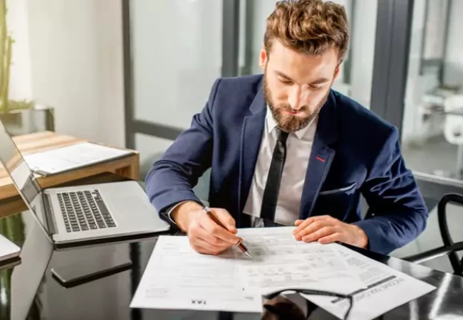 A person in a suit and tie sitting at a desk with a laptop and papers